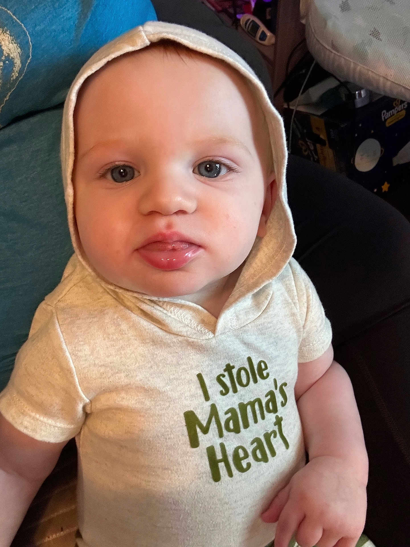 Elias is registered to the contest to win money with this photo: baby, child, hoodie, blue_eyes, indoor, cute, infant, shirt, text_on_clothing, portrait, person, sitting, young_child, face, expression, casual_clothing, hand, apparel, closeup, soft_lighting