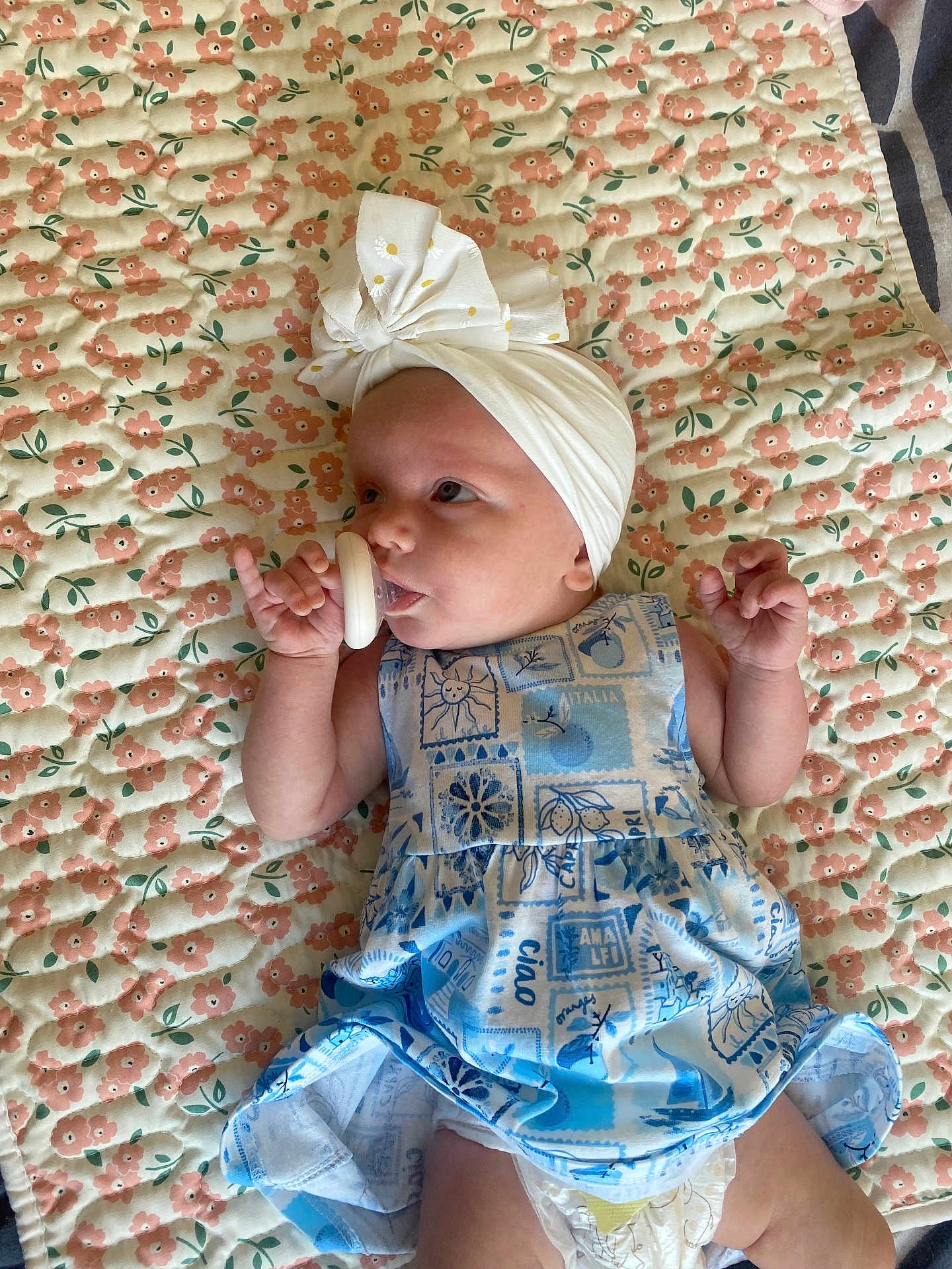 Poppy Rae is registered to the contest to win money with this photo: baby, pacifier, headwrap, bow, dress, floral_quilt, patterned_clothing, infant, lying_down, diaper, cute, indoor, child, small_hand, soft_lighting, portrait, fabric, pink_flowers, blue_dress, relaxed