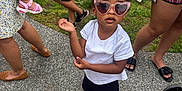 Sayora is registered to the contest to win money with this photo: toddler, child, sunglasses, heart_shaped, pink_bow, white_tshirt, black_shorts, sneakers, outdoor, grass, pavement, people, stroller, fence, casual_pose, summer_clothing, legs, footwear, accessories, daytime