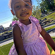 Sayora is registered to the contest to win money with this photo: child, girl, smiling, outdoor, sunlight, dress, purple_dress, playground, grass, fence, tree, daylight, sunflare, happy, portrait, person, curly_hair, earrings, summer, fun
