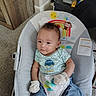 baby, infant, baby_rocker, mittens, blue_clothing, car_print, indoor, carpet, wooden_furniture, cute, child, seated, colorful, toy_design, smiling, face, person, comfortable, home, curious