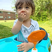 Jasmin is registered to the contest to win money with this photo: toddler, child, baby, walker, outdoor, grass, trees, greenery, sunny, daylight, curly_hair, blue, orange, toy, snack, face, person, expression, nature, casual_clothing