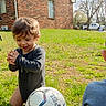 toddler, child, soccer_ball, grass, outdoor, brick_house, sunny, playing, kneeling, happy, clapping, young_child, daylight, casual_clothing, nature, greenery, smiling, person, recreation, sport