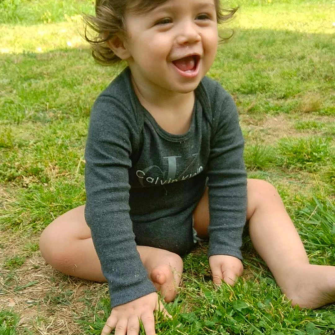 Jasmin is registered to the contest to win money with this photo: toddler, child, smiling, grass, outdoor, curly_hair, happy, nature, baby, sitting, greenery, playful, daylight, casual_clothing, person, young_child, fun, joy, barefoot, laughter