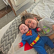 Albie is registered to the contest to win money with this photo: baby, child, superman_costume, smiling, bed, blanket, sibling, happy, cute, indoor, family, young_child, playing, joyful, clothing, portrait, face, toddler, cuddling, resting