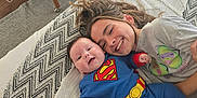 Albie is registered to the contest to win money with this photo: baby, child, superman_costume, smiling, bed, blanket, sibling, happy, cute, indoor, family, young_child, playing, joyful, clothing, portrait, face, toddler, cuddling, resting