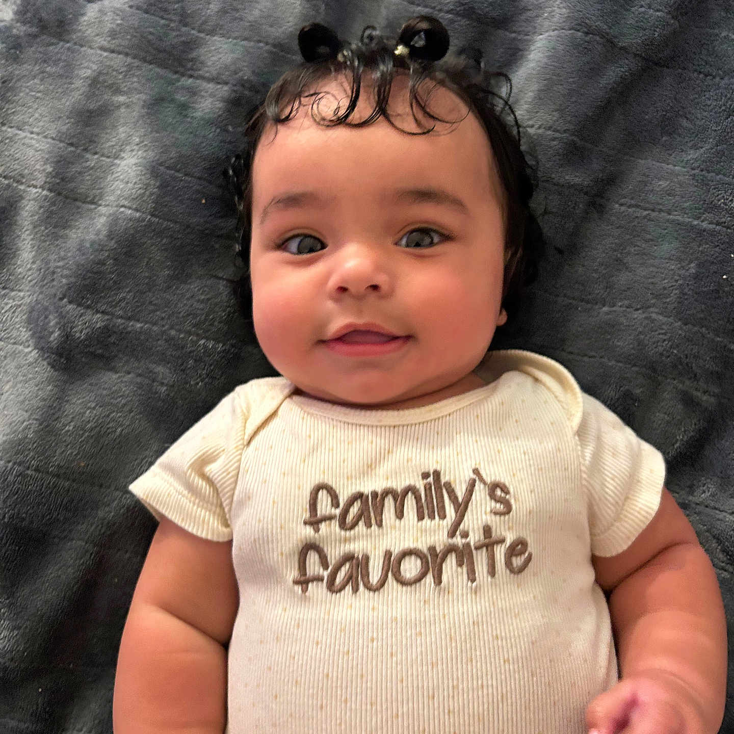 Levaeh joined the competition — help win amazing prizes! baby, infant, cute, smiling, onesie, clothing, curly_hair, hair_buns, blanket, soft_texture, face, portrait, child, lying_down, indoors, happy, chubby_cheeks, hand, person, family