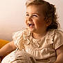 toddler, child, smiling, laughing, curly_hair, cream_dress, crochet, floral, ball, patterned_ball, sitting, happy, indoor, soft_lighting, portrait, cute, baby, face, hand, joy