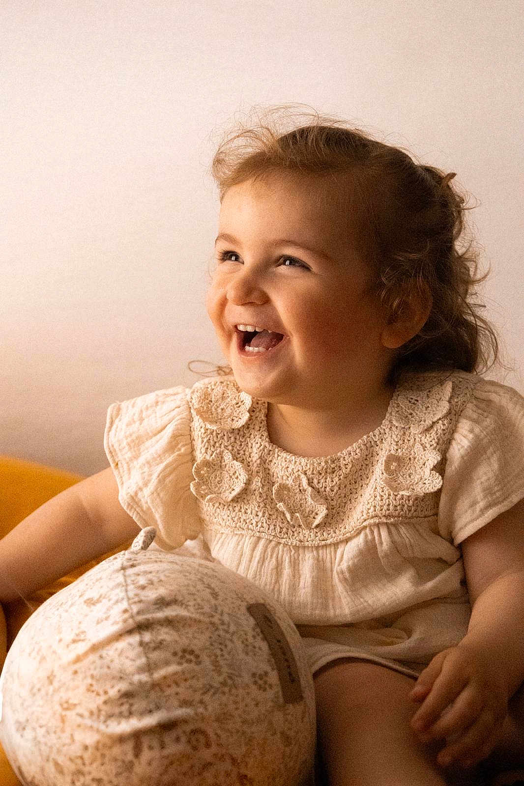 Louise participe au concours pour gagner de l'argent avec cette photo : toddler, child, smiling, laughing, curly_hair, cream_dress, crochet, floral, ball, patterned_ball, sitting, happy, indoor, soft_lighting, portrait, cute, baby, face, hand, joy