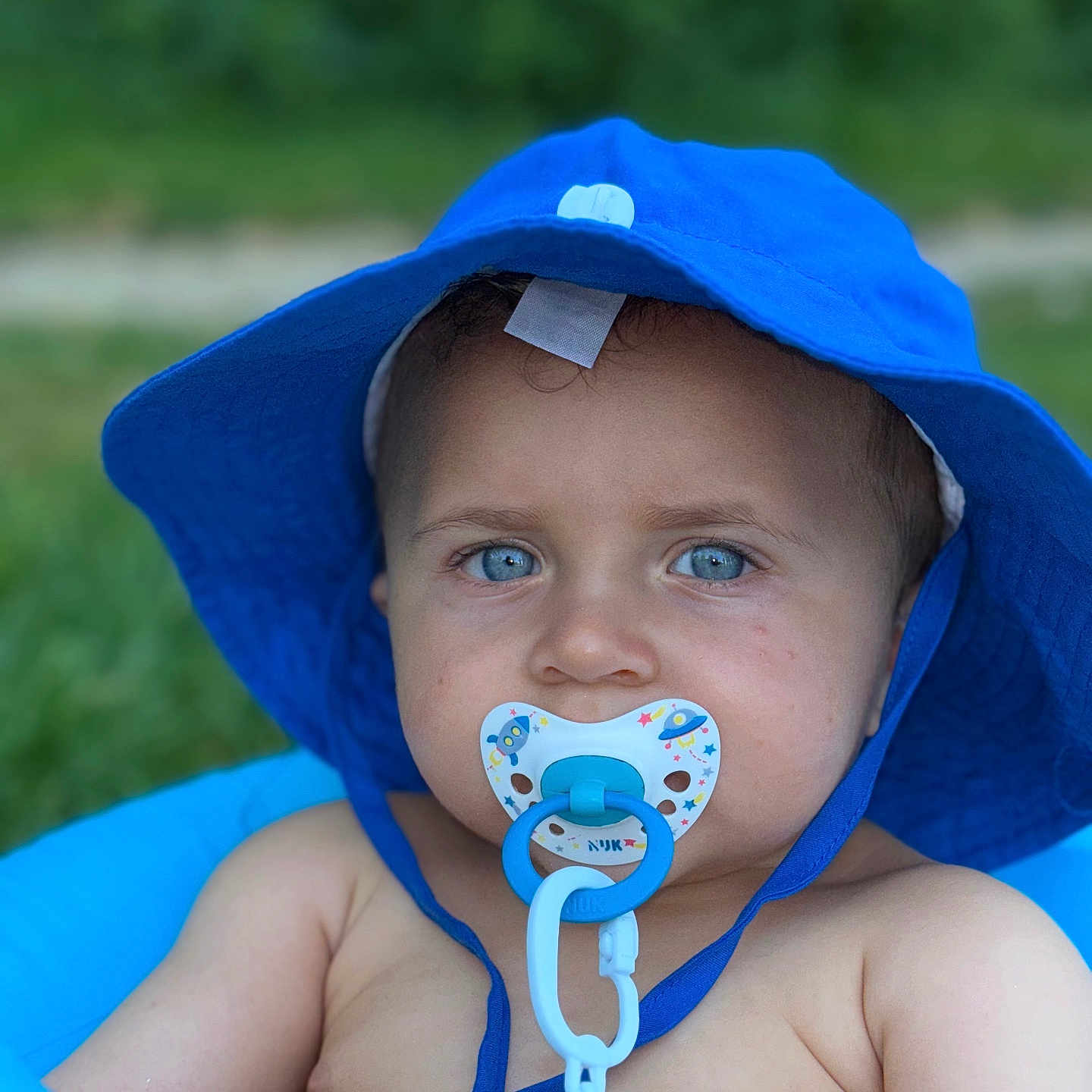 Seydou a rejoint le concours — aidez-le/la à gagner de superbes lots ! baby, baby_boy, baby_toy, blue_eyes, blue_hat, chained_pacifier, child, close_up, cute, daylight, face, green_background, infant, outdoor, pacifier, portrait, skin, soft_focus, summer, sun_hat