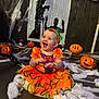 baby, carved_pumpkins, celebration, child, cobwebs, cute, decorations, festive, halloween, happy, headband, holiday, indoor, orange_dress, person, pumpkin_costume, shadow, sitting, smiling, wall