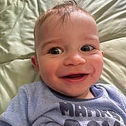 Josiah is registered to the contest to win money with this photo: baby, smiling, face, blue_shirt, text_on_clothing, lying_down, quilted_surface, close_up, child, happy, infant, cute, portrait, young_child, head, skin, hair, eyes, mouth, person