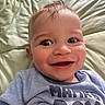 baby, smiling, face, blue_shirt, text_on_clothing, lying_down, quilted_surface, close_up, child, happy, infant, cute, portrait, young_child, head, skin, hair, eyes, mouth, person