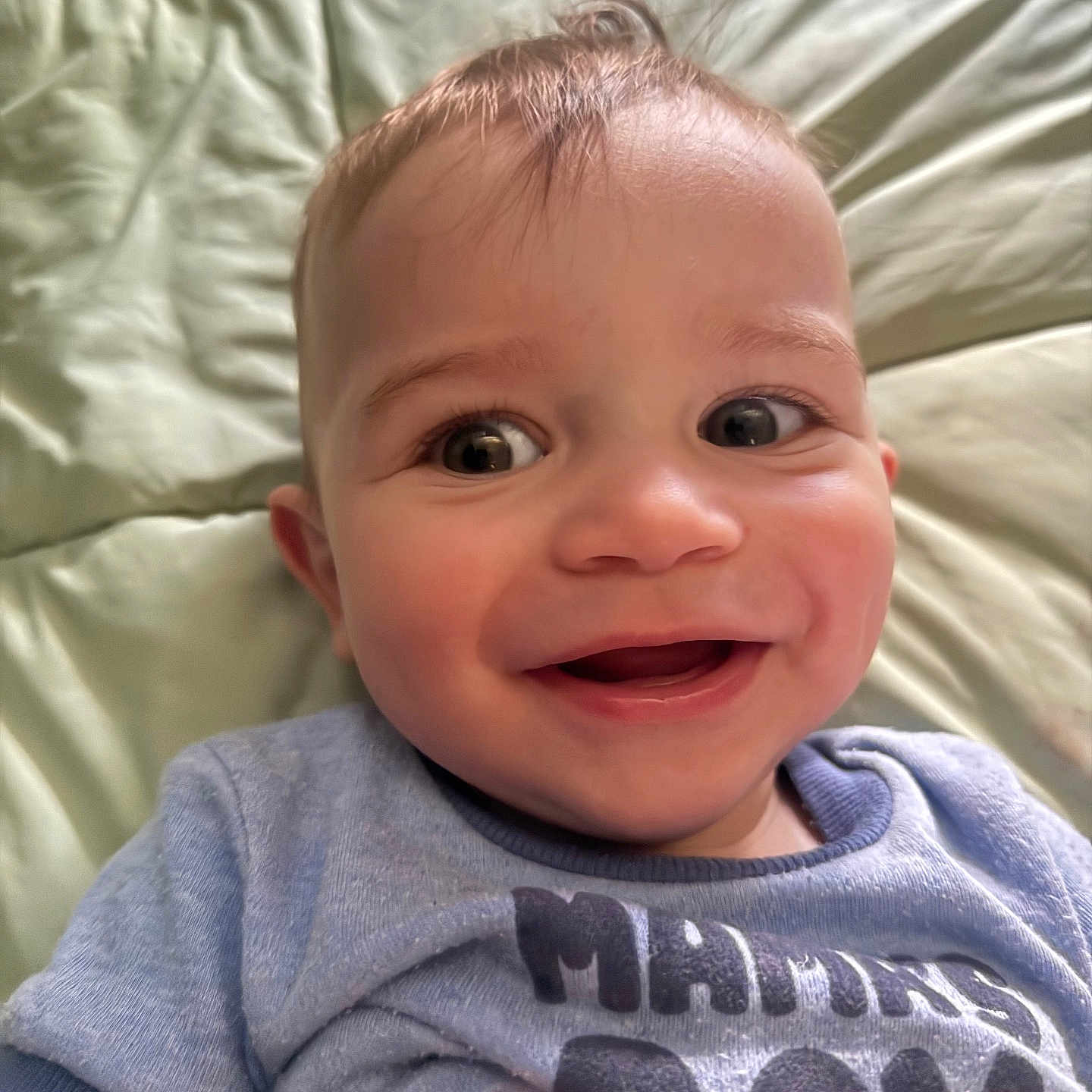 Josiah is registered to the contest to win money with this photo: baby, smiling, face, blue_shirt, text_on_clothing, lying_down, quilted_surface, close_up, child, happy, infant, cute, portrait, young_child, head, skin, hair, eyes, mouth, person