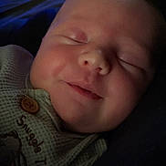 Kylee is registered to the contest to win money with this photo: baby, bed, blanket, blue, button, close_up, clothing, cozy, dim_lighting, face, head, indoors, infant, knitwear, newborn, peaceful, portrait, resting, sleeping, smiling