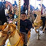 child, carousel, horse, amusement_park, ride, smiling, jacket, people, fun, family, entertainment, golden_pole, indoor, happy, toy, playful, casual_clothing, background, colorful, crowd