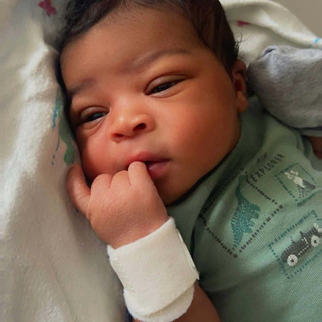 Ahmiri joined the competition — help win amazing prizes! newborn, baby, infant, child, portrait, close_up, face, eyes, smiling, hand, fist, blanket, swaddled, green_outfit, clothing, lying_down, indoors, bed, soft_lighting, photography