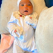 Maryam is registered to the contest to win money with this photo: baby, infant, white_outfit, embroidery, blanket, couch, soft_texture, headwear, mittens, surprised_expression, lying_down, indoor, person, child, cute, comfortable, plush, fabric, home, portrait