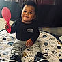 Zamari is registered to the contest to win money with this photo: toddler, child, playful, smiling, indoor, blanket, space_theme, curly_hair, black_shirt, grey_pants, black_sneakers, toy, pink_paddle, bed, curtains, home, portrait, cute, happy, sitting