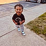 Zamari is registered to the contest to win money with this photo: toddler, child, smiling, sidewalk, sneakers, curly_hair, black_tshirt, outdoor, daylight, grass, car, parking_lot, happy, walking, person, cute, casual_clothing, urban, young_child, joyful