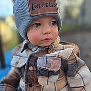 Lincoln is registered to the contest to win money with this photo: toddler, child, hat, beanie, jacket, plaid, face, portrait, outdoor, cold_weather, young_child, curious, warm_clothing, button, eyewear, closeup, person, cute, soft_focus, winter