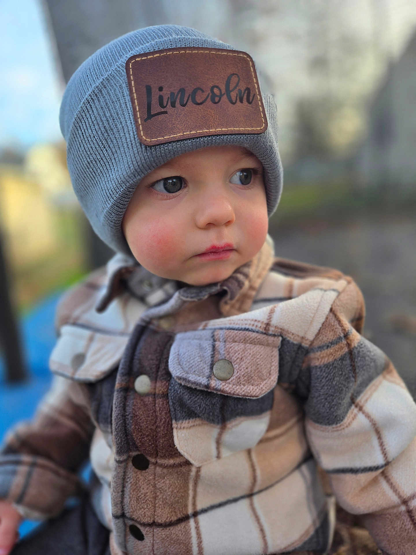 Lincoln is registered to the contest to win money with this photo: toddler, child, hat, beanie, jacket, plaid, face, portrait, outdoor, cold_weather, young_child, curious, warm_clothing, button, eyewear, closeup, person, cute, soft_focus, winter