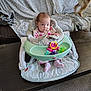 baby, infant, baby_chair, toy, pink_clothing, polka_dot, face, expression, couch, blanket, dog_tail, dog_leg, carpet, wooden_table, indoor, home, furniture, person, child, seated