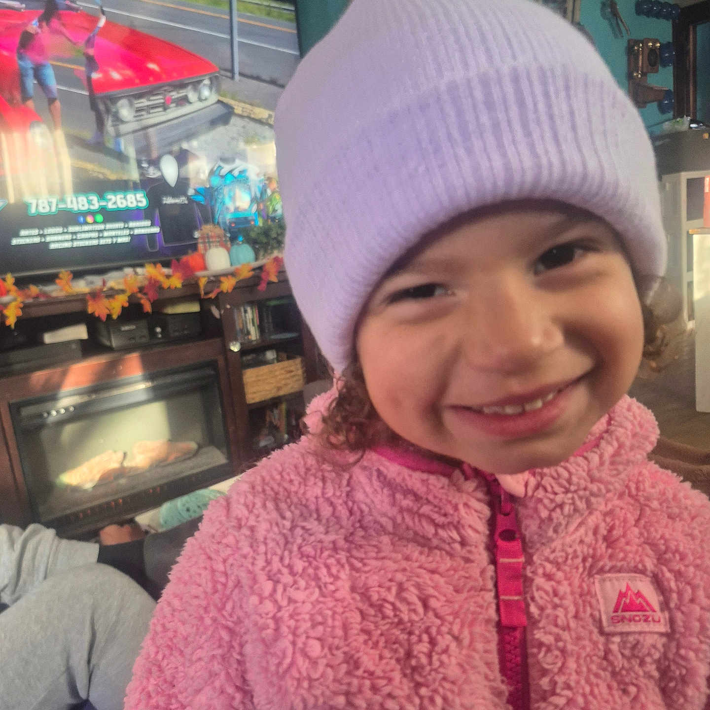 Arianelys Otero joined the competition — help win amazing prizes! autumn_decor, beanie, casual, child, colorful, cozy, curly_hair, entertainment, face, furniture, happy, home, indoor, living_room, person, pink_jacket, portrait, smile, television, warm_clothing