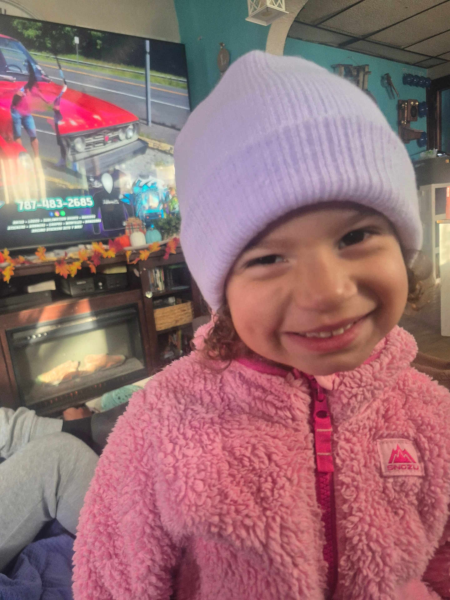 Arianelys Otero joined the competition — help win amazing prizes! child, smile, pink_jacket, beanie, indoor, living_room, television, autumn_decor, furniture, cozy, curly_hair, face, person, warm_clothing, casual, home, entertainment, colorful, happy, portrait