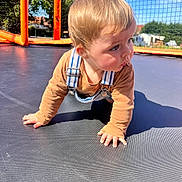 Alessio participe au concours pour gagner de l'argent avec cette photo : toddler, child, crawling, trampoline, outdoor, net, orange, blue_sky, sunlight, tree, grass, clothing, overalls, long_sleeve, face, hands, shadow, daytime, play, curious
