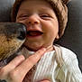 child, baby, infant, smiling, knit_hat, brown_hat, dog_nose, dog, hand, tattoo, onesie, white_clothing, closeup, portrait, indoor, couch, table, water_bottle, laughter, pet_interaction