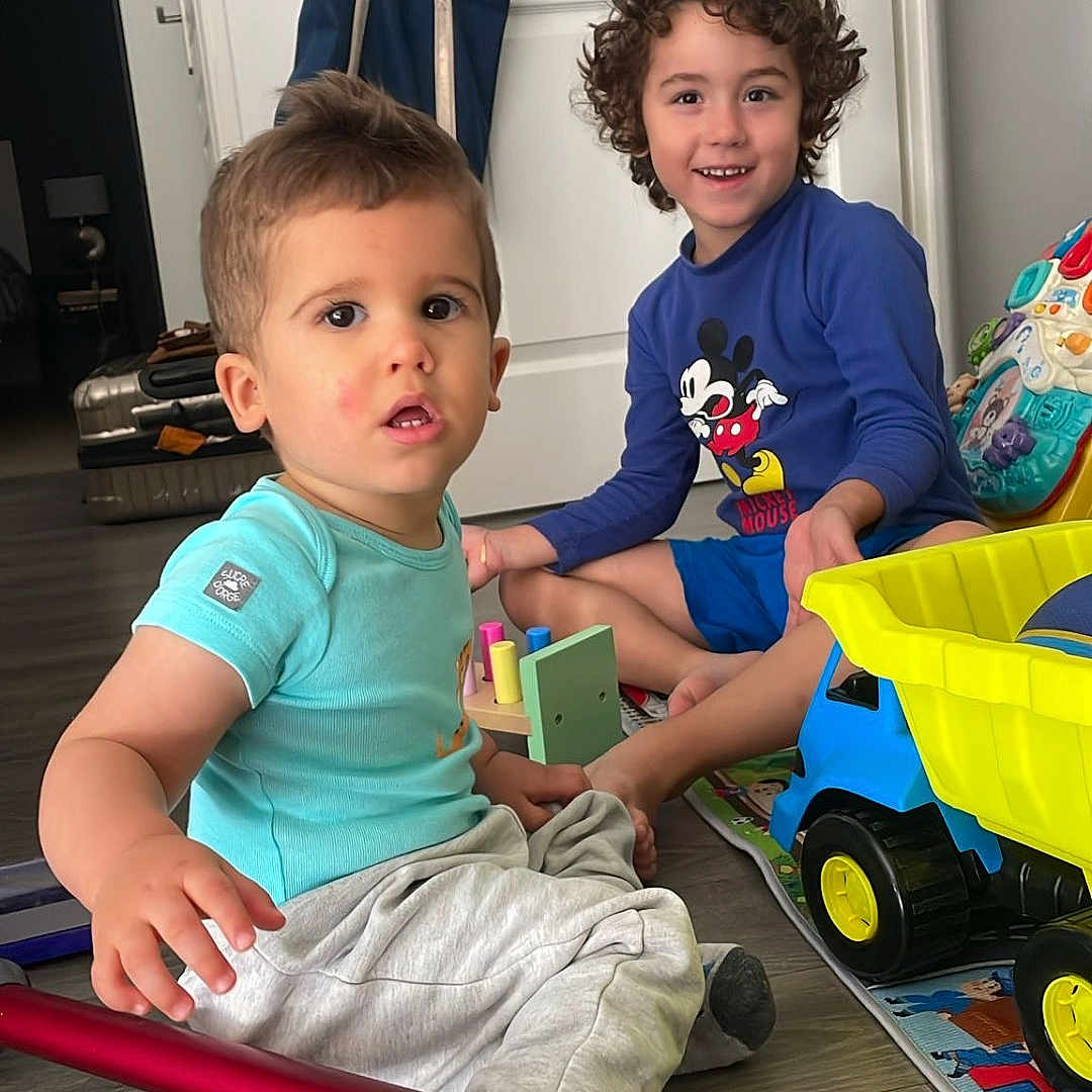 Enzo Meireles a rejoint le concours — aidez-le/la à gagner de superbes lots ! boy, candid, casual_clothing, child, children, colorful, curious_expression, curly_hair, floor, home, indoor, mickey_mouse_shirt, playtime, sitting, smiling, toy_truck, toys, vacuum_cleaner, wooden_floor, young_child