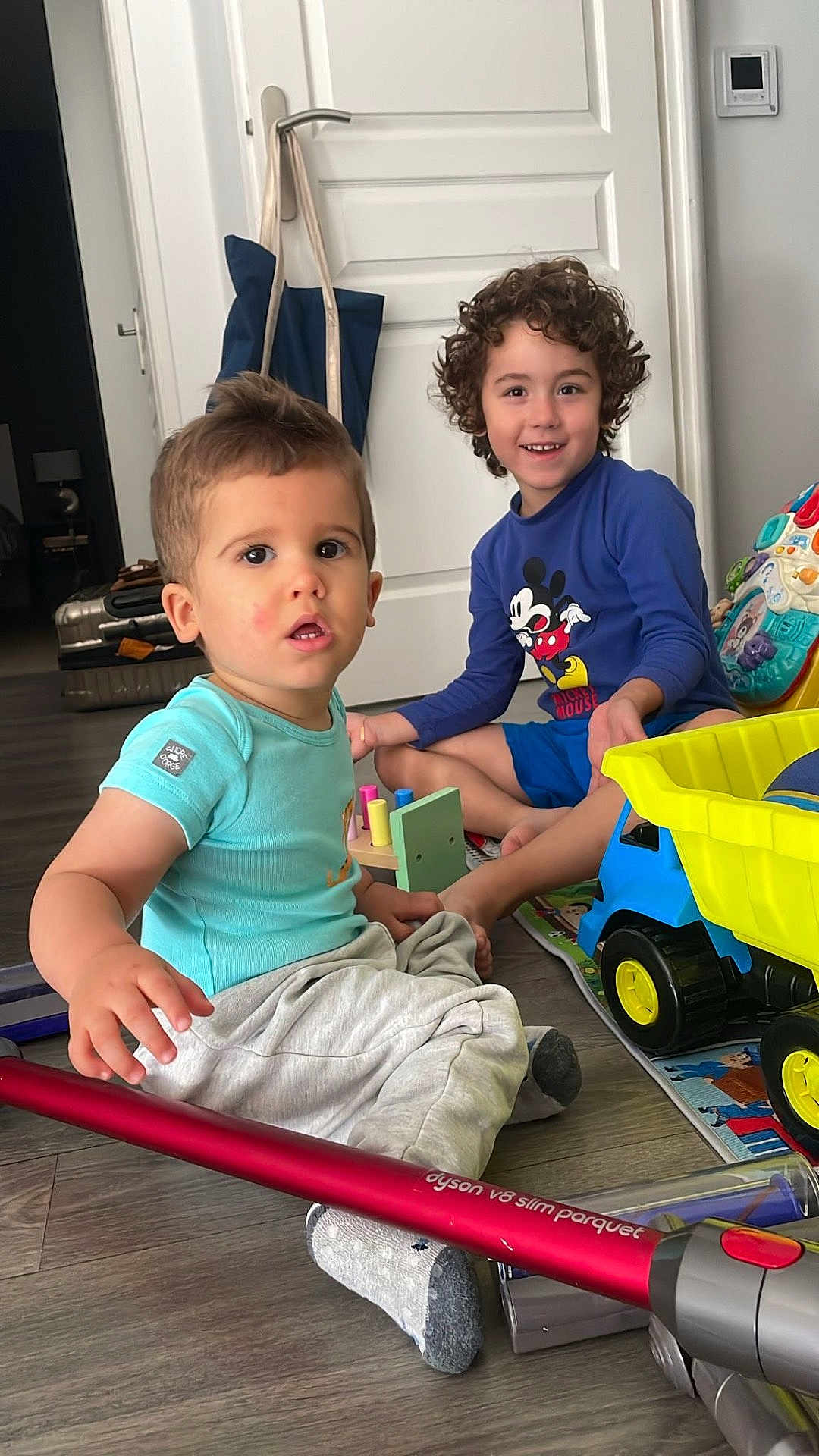 Enzo Meireles a rejoint le concours — aidez-le/la à gagner de superbes lots ! boy, candid, casual_clothing, child, children, colorful, curious_expression, curly_hair, floor, home, indoor, mickey_mouse_shirt, playtime, sitting, smiling, toy_truck, toys, vacuum_cleaner, wooden_floor, young_child
