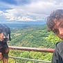 person, dog, outdoor, nature, landscape, greenery, curly_hair, railing, sky, clouds, valley, holding, pet, casual_clothing, portrait, animal, scenic, daylight, leisure, travel