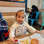 Chloe joined the competition — help win amazing prizes! child, smile, hoodie, food, burger, french_fries, ketchup, table, restaurant, person, chair, indoor, casual_clothing, happy, face, hair, wood, napkin, seat, fast_food