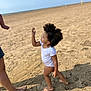 child, toddler, beach, sand, barefoot, afro_hair, white_clothing, person, outdoor, sunny, shadow, summer, reaching, curious, small_child, standing, adult_leg, nature, sky, playful