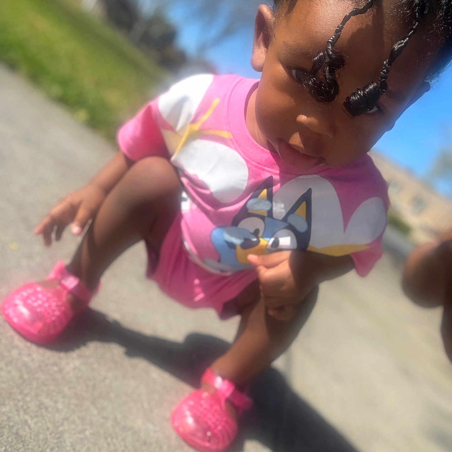 Love is registered to the contest to win money with this photo: child, toddler, pink_shoes, cartoon_tshirt, outdoor, sunny, sidewalk, curly_hair, squatting, playful, face, person, daylight, casual_clothing, young_child, skin, expression, shadow, grass, background