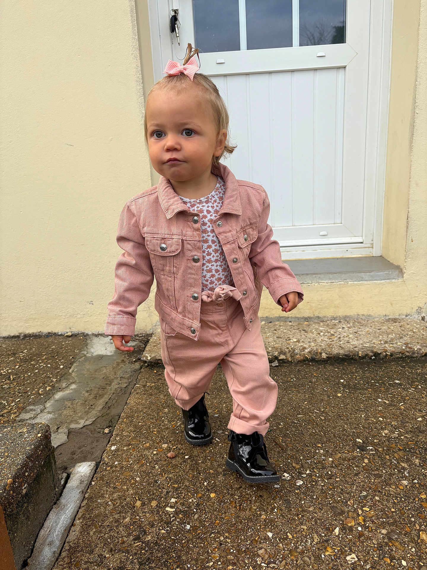 Julia participe au concours pour gagner de l'argent avec cette photo : toddler, child, pink_jacket, pink_pants, black_boots, bow, hair_accessory, outdoor, pavement, door, concrete, sidewalk, walking, serious_expression, young_child, person, clothing, footwear, curly_hair, casual