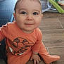 baby, child, infant, orange_clothing, floor, sitting, smiling, cute, indoors, wooden_floor, person, young_child, long_sleeve, footed_pajamas, face, eyes, hand, head, expression, home