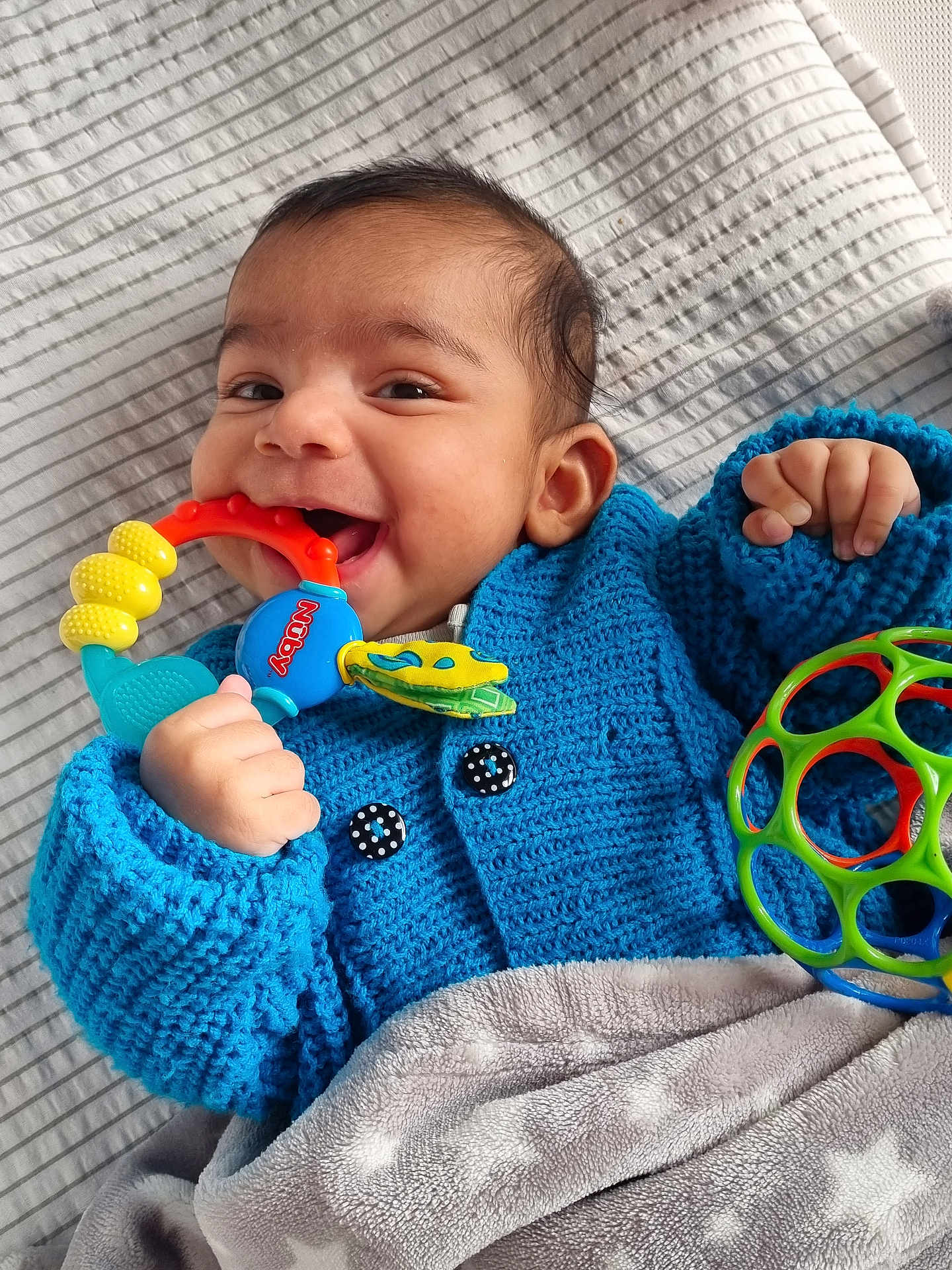 Shazil is registered to the contest to win money with this photo: baby, smiling, blue_sweater, teething_toy, plastic_ball, blanket, lying_down, happy, infant, cute, child, knitted_clothing, indoors, playful, hand, face, head, portrait, young_child, soft_texture