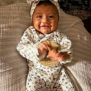Rebekah joined the competition — help win amazing prizes! baby, child, infant, smiling, happy, portrait, lying_down, bed, bedroom, pajamas, onesie, headband, bow, hello_kitty, patterned_clothing, hands, feet, blanket, indoor, cute