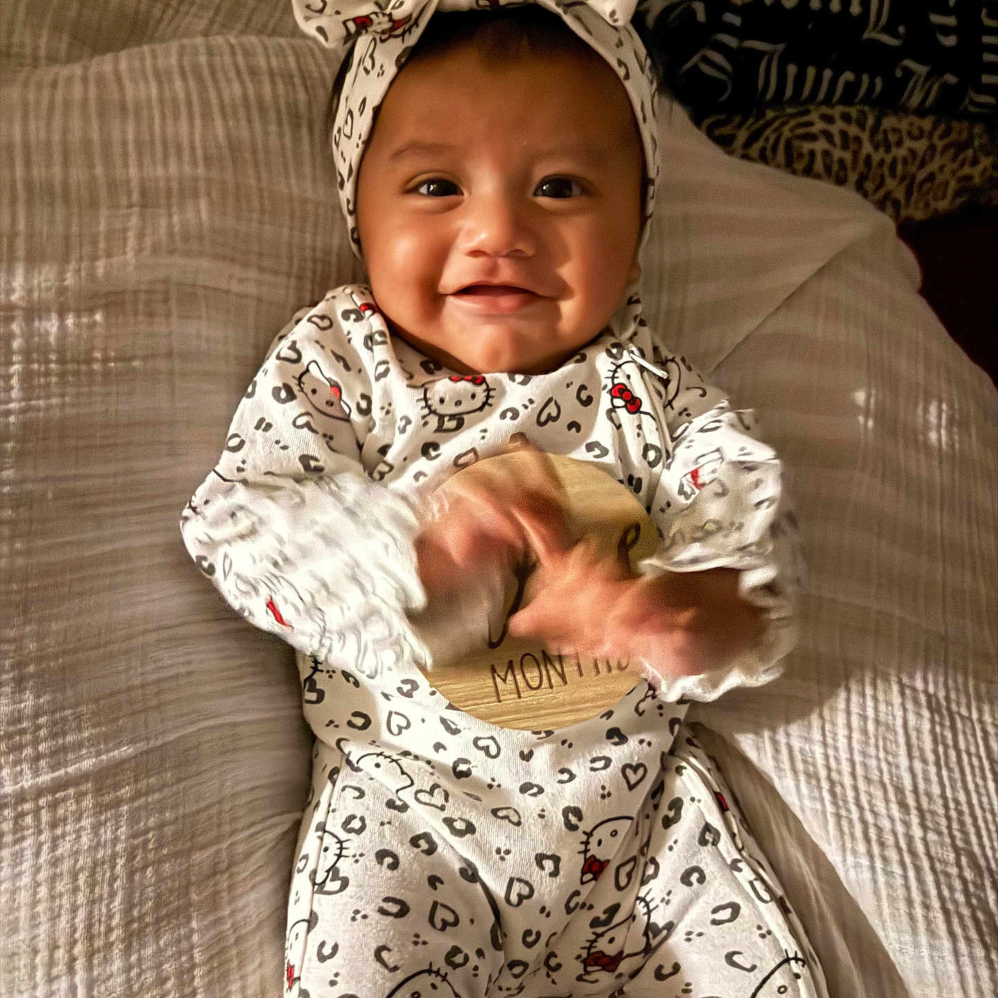 Rebekah joined the competition — help win amazing prizes! baby, child, infant, smiling, happy, portrait, lying_down, bed, bedroom, pajamas, onesie, headband, bow, hello_kitty, patterned_clothing, hands, feet, blanket, indoor, cute