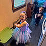 child, toddler, girl, dress, tutu, smile, indoor, floor, window, toy, party, headband, hair, clothing, person, furniture, table, chair, black_shirt, happy