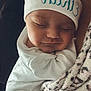baby, sleeping, newborn, beanie, white_clothing, blanket, leopard_print_blanket, closeup, face, eyes_closed, peaceful, cozy, parent_arm, skin, portrait, indoor, nap, soft_texture, headwear, gentle_lighting