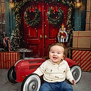 Hugh is registered to the contest to win money with this photo: baby, toy_car, smiling, festive, christmas, wreath, presents, holiday, nutcracker, red_door, indoor, child, jeans, sweater, boots, floor, decorations, seasonal, happy, portrait