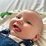 baby, cheeks, child, close_up, clothing, cute, denim_overalls, ear, eyes, face, happy, head, indoor, infant, person, skin, smiling, soft_background, tongue, white_shirt