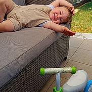 Ismaël participe au concours pour gagner de l'argent avec cette photo : child, toddler, smiling, sofa, wicker, outdoor, patio, toy, tricycle, green, blue, beige_clothing, happy, relaxed, sunlight, fence, grass, trees, daylight, play