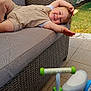 child, toddler, smiling, sofa, wicker, outdoor, patio, toy, tricycle, green, blue, beige_clothing, happy, relaxed, sunlight, fence, grass, trees, daylight, play