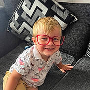Leo is registered to the contest to win money with this photo: child, boy, glasses, red_glasses, smiling, couch, cushion, remote_control, shorts, shirt, indoor, blond_hair, happy, sitting, home, furniture, portrait, casual_clothing, playful, young