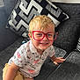 child, boy, glasses, red_glasses, smiling, couch, cushion, remote_control, shorts, shirt, indoor, blond_hair, happy, sitting, home, furniture, portrait, casual_clothing, playful, young